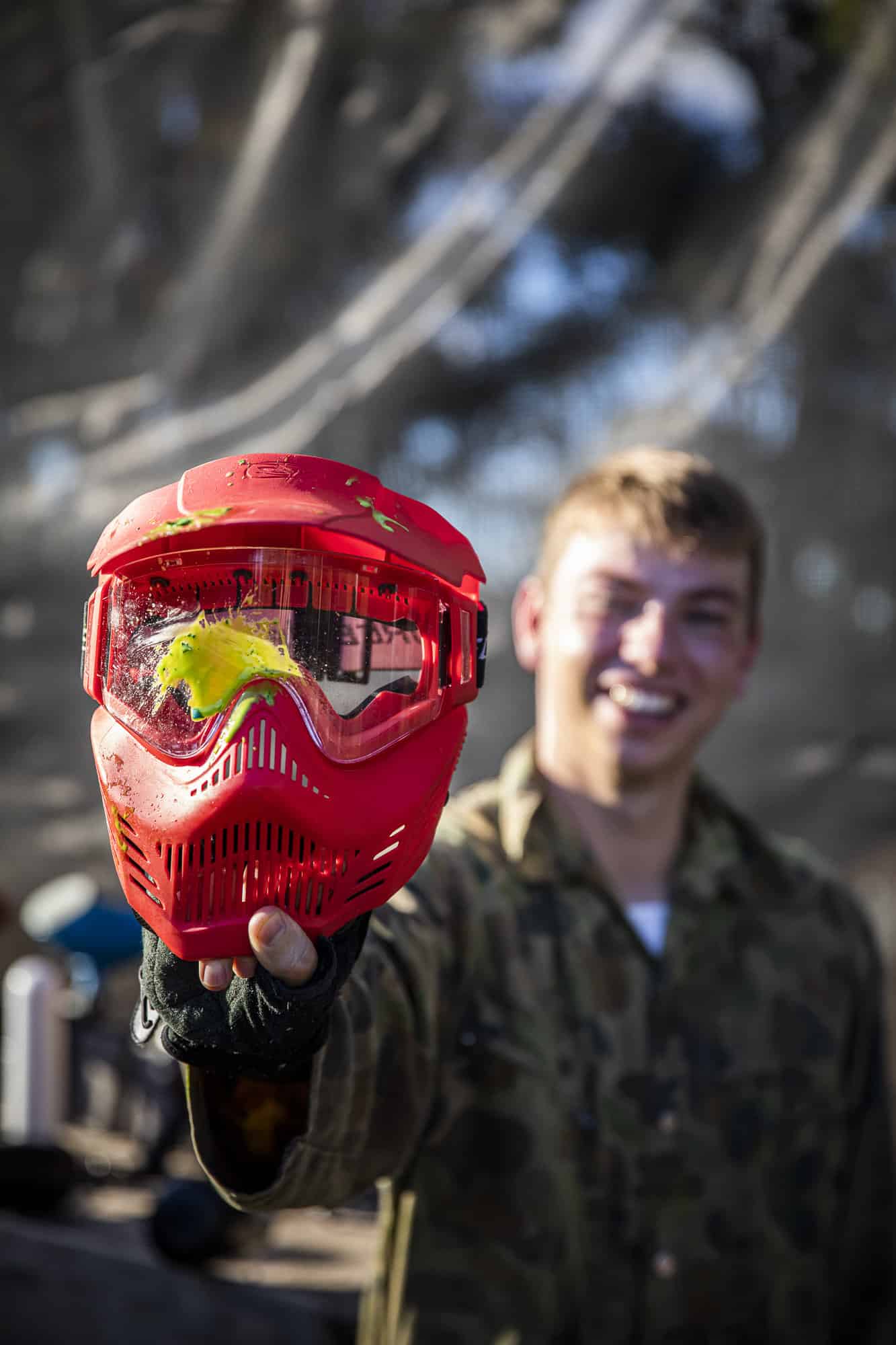 A young man holding up a paintball helmet with a paint splatter